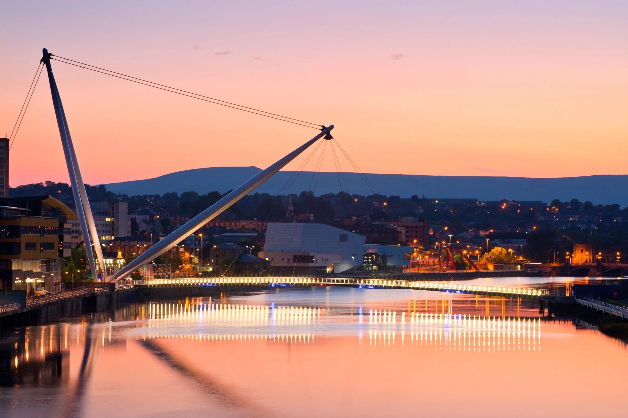 Cityscape at dusk with a large modern bridge over a river, reflected in the water, and a mountain range in the background. The sky has pink and purple hues with some clouds.