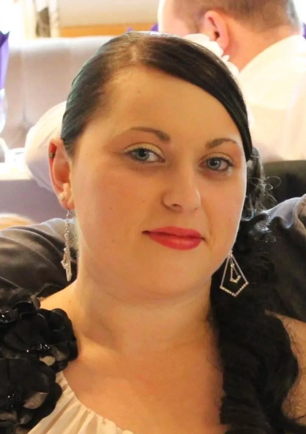 Close-up of a woman with dark hair, light blue eyes, red lipstick, wearing earrings and a dress with black ruffle details on the shoulders, smiling at the camera in a formal setting.