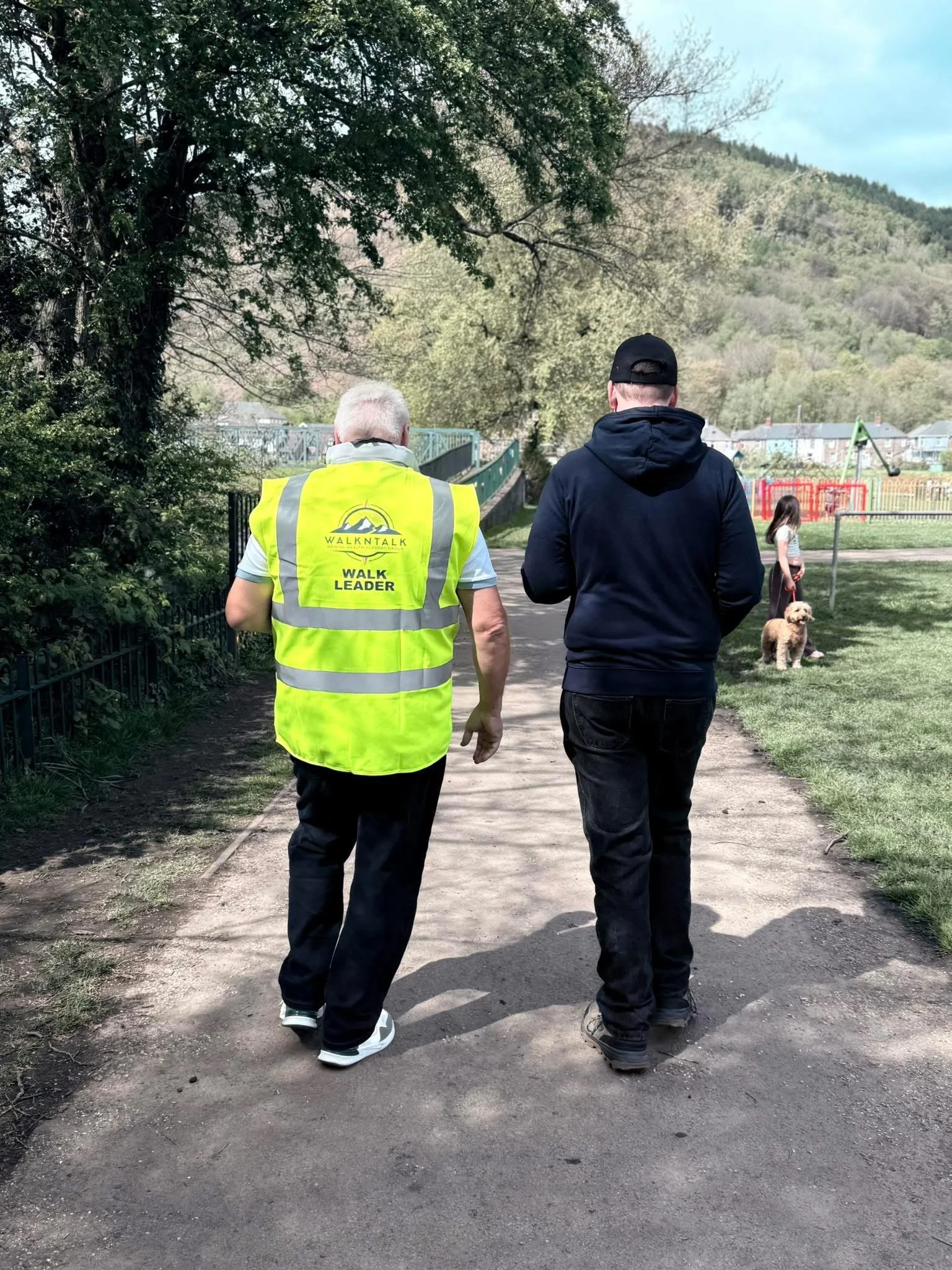 Two men walking on a paved path in a park, one wearing a bright yellow safety vest with 'Walk Leader' printed on the back, and the other in dark clothing and a black cap. In the background, a young girl with a dog stands near a fence and playground equipment, with trees and hills in the distance.