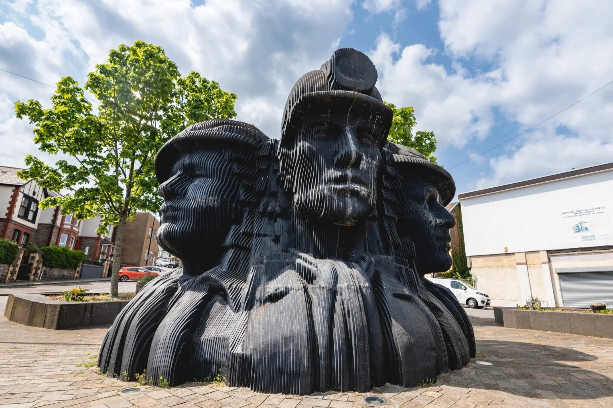 Sculpture of three black interlocked heads with stylized lines, set outdoors on a brick plaza, with green trees and a partly cloudy sky in the background.