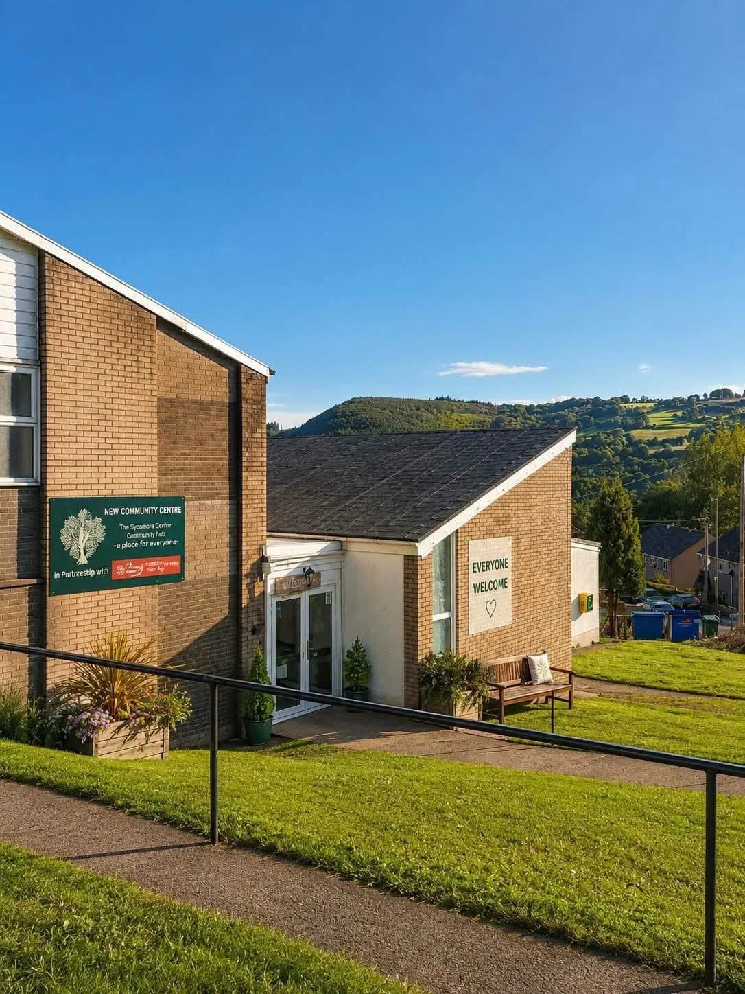 A community center building with a green sign and a welcoming sign that says 'Everyone Welcome'. The building is surrounded by a grassy lawn and has flowers and plants near the entrance. Hills and houses are visible in the background under a blue sky.