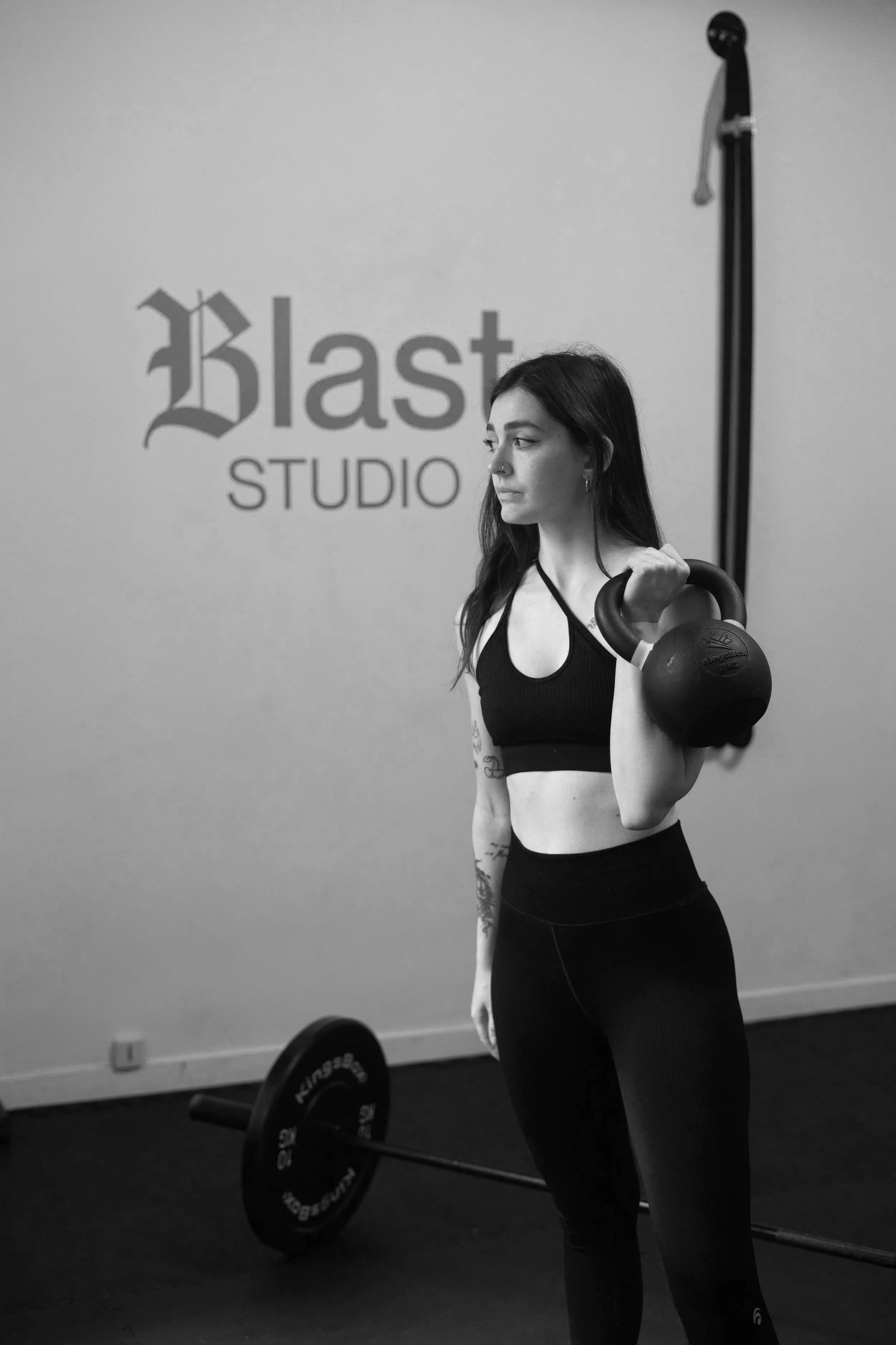 Une femme portant une tenue de sport, tient une kettlebell dans la main lors d'une séance d'entraînement dans un studio de fitness nommé 'Blast Studio'.