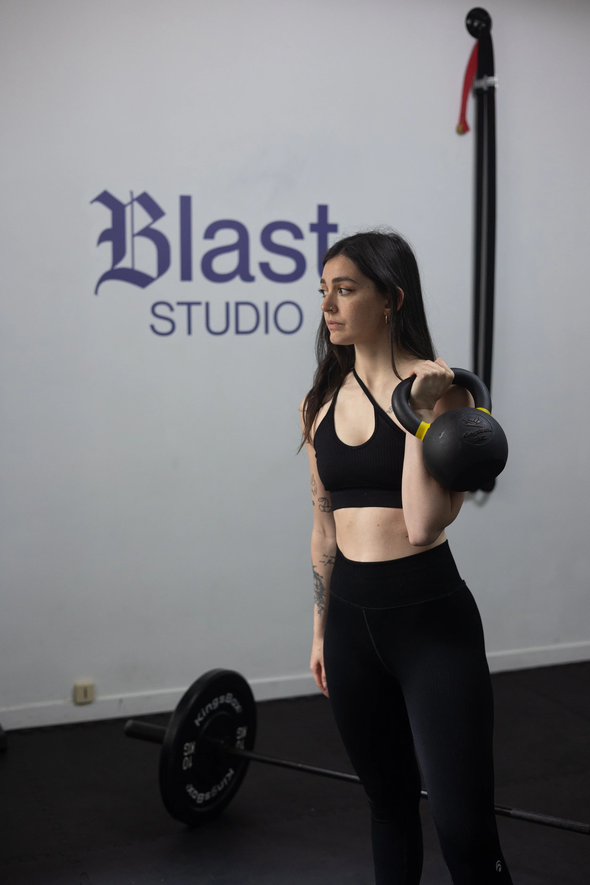 Une femme debout, en tenue de sport, dans une salle de sport, tenant une kettlebell dans sa main droite. Le fond affiche le logo 'Blast Studio' sur un mur blanc, et il y a une barre de poids au sol.