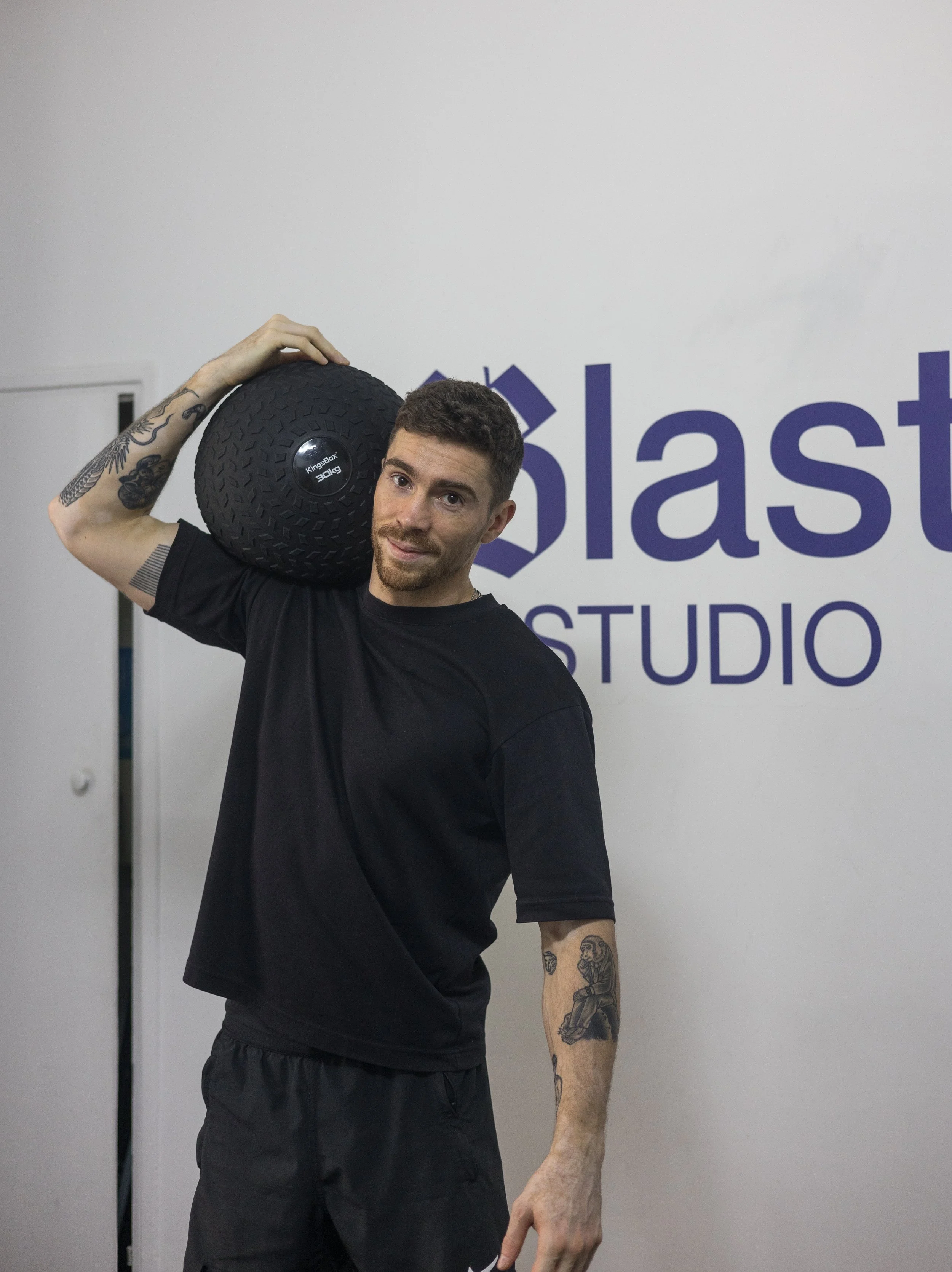 Un homme musclé porte un poids sur son épaule dans une salle de sport, avec un mur derrière lui portant le logo 'Blast Studio'.