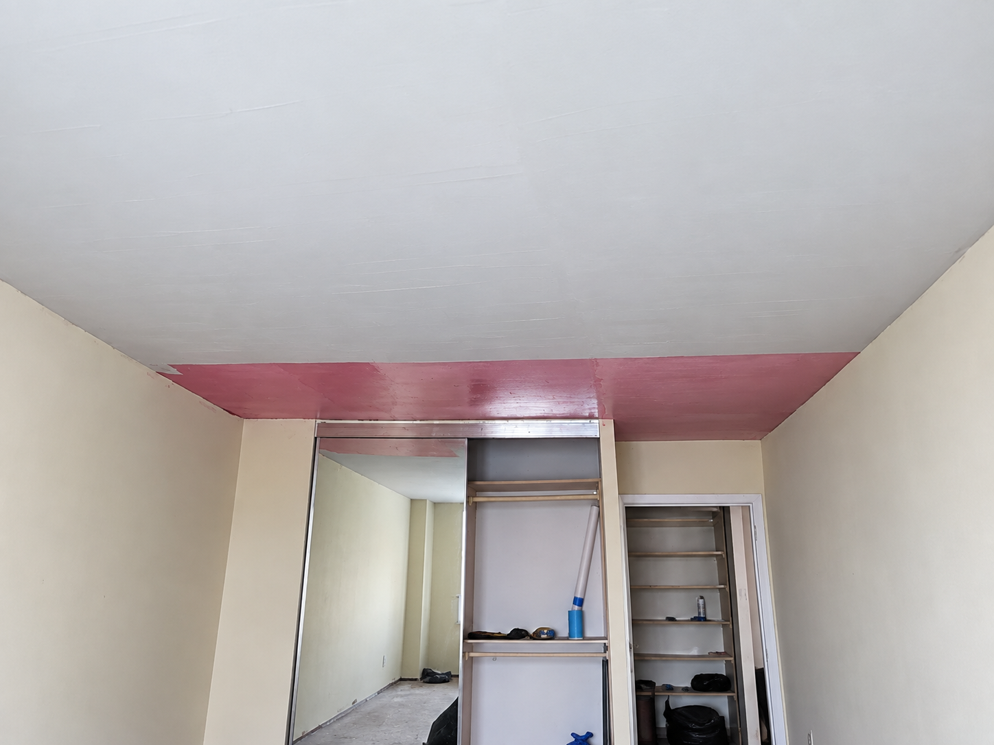 Sala en proceso de construcción o renovación, con paredes claras, techo pintado parcialmente en rojo y estructura de pared con estanterías y herramientas.