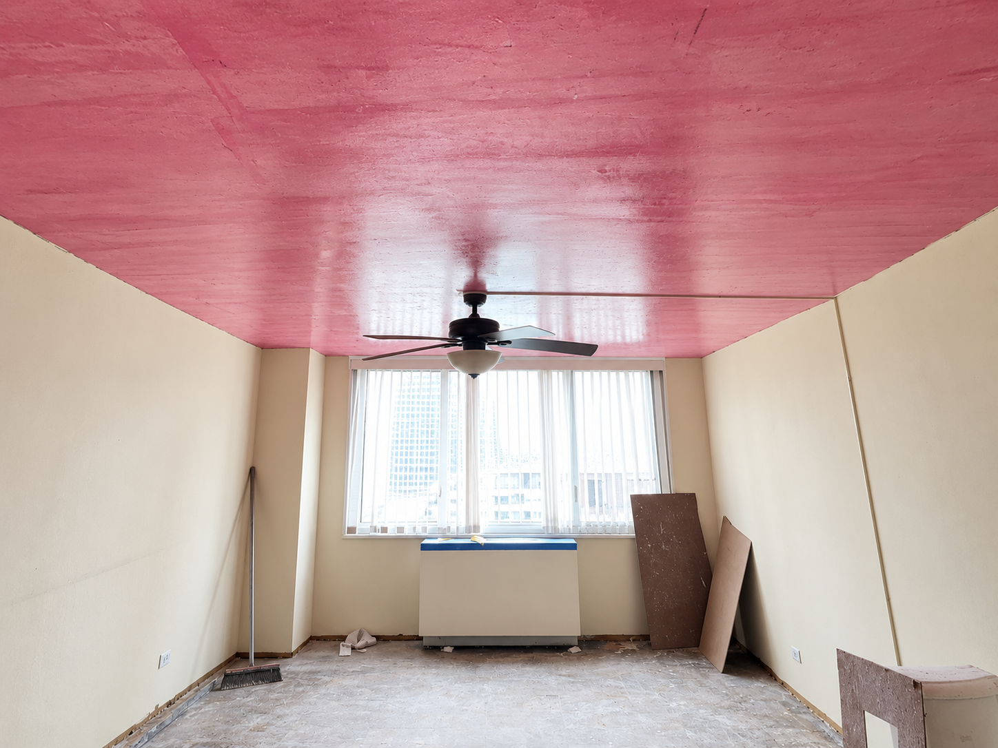 Sala en remodelación con paredes amarillas, techo rosa y ventilador de techo, ventanas con cortinas blancas y suelo en proceso de reparación.