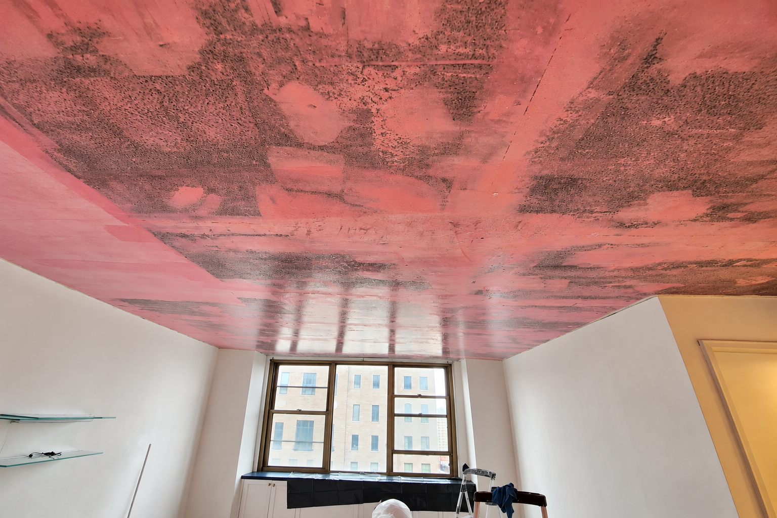 Techo en proceso de pintura con color rosa y detalles en negro, habitación con ventanas grandes, paredes blancas y escaleras, en un espacio interior.
