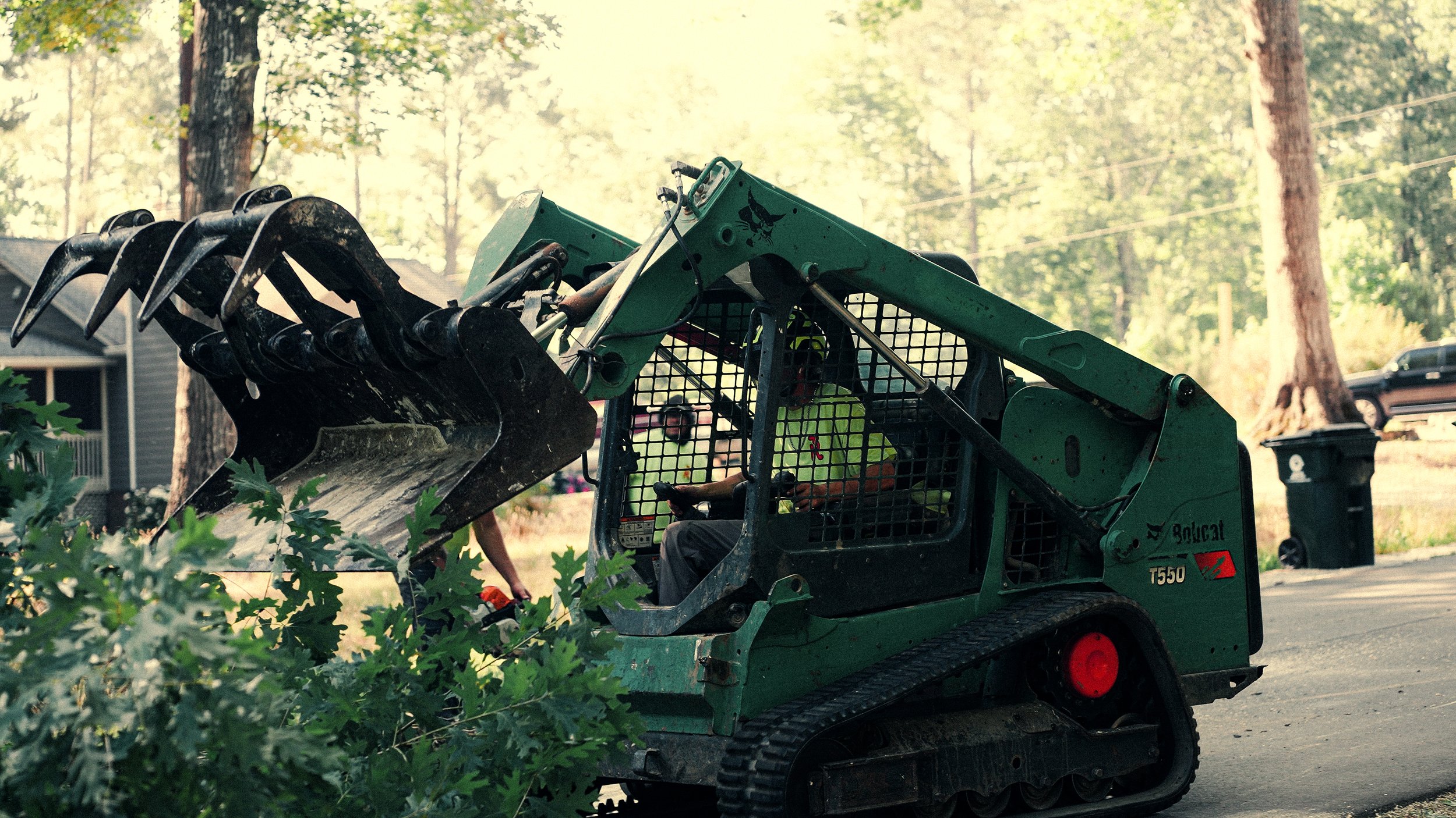 A person operating a small green Bobcat T550 skid-steer loader with a large black bucket attachment, clearing or moving greenery in a residential neighborhood surrounded by trees.