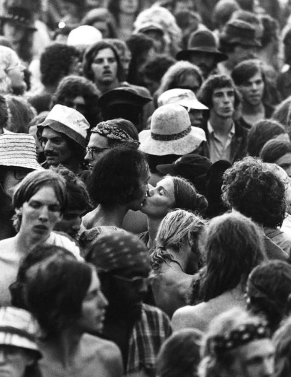 Black and white photo of a large crowd of women, some wearing hats, with a couple kissing in the center.