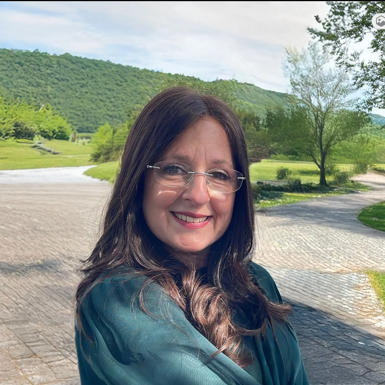 A woman with long brown hair and glasses smiling outdoors in front of a park with green trees, a grassy field, and mountains in the background.