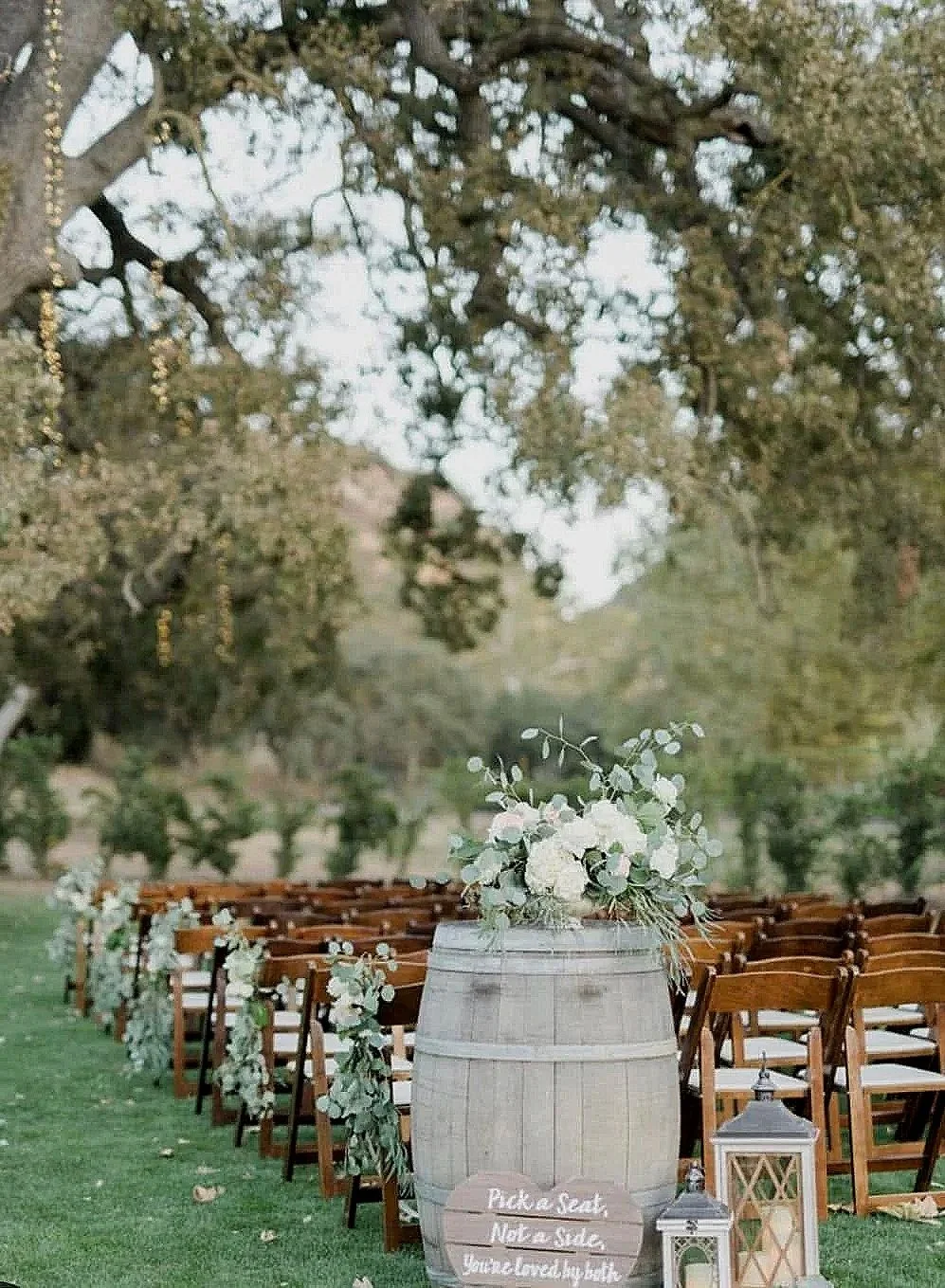 Outdoor wedding ceremony setup with wooden chairs decorated with white flowers, a large barrel with a floral arrangement, and lanterns nearby. Trees and greenery in the background.