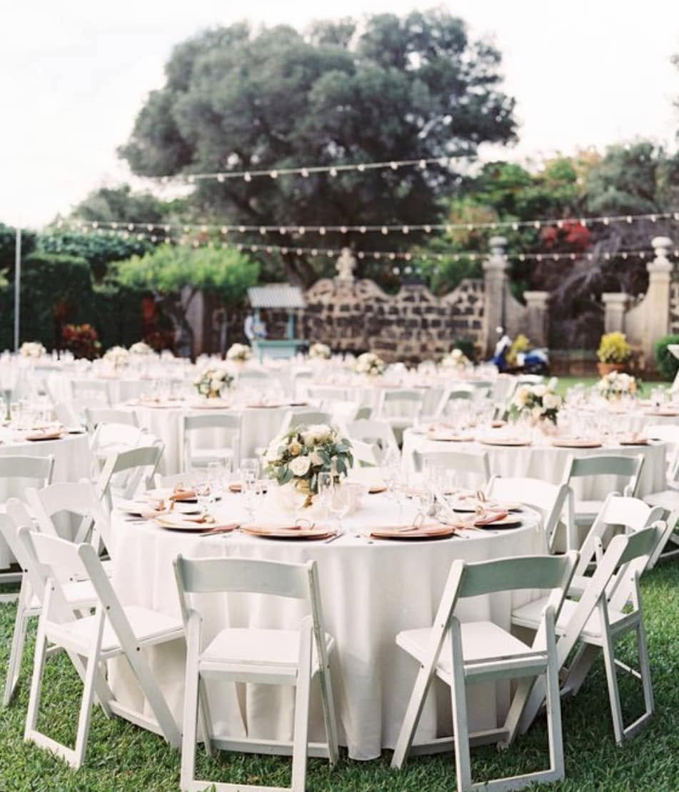 Outdoor event setup with round tables covered in white tablecloths, decorated with floral centerpieces, surrounded by white chairs, with string lights hanging above and a large tree in the background.