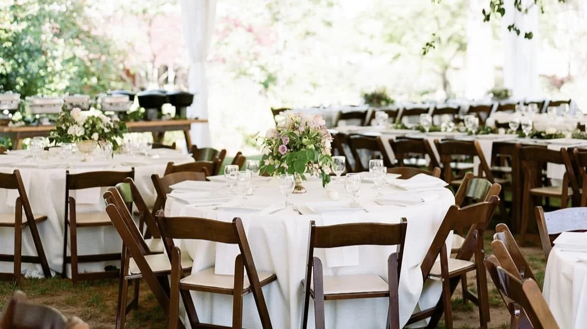 Wedding reception setup with round tables covered in white tablecloths, decorated with floral centerpieces, set with glassware, and surrounded by wooden chairs, in a bright outdoor setting.