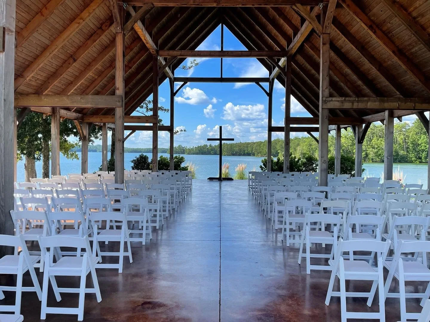 An outdoor lakeside wedding venue with wooden structure, white chairs, a cross at the end, and a scenic view of water and trees under a partly cloudy sky.
