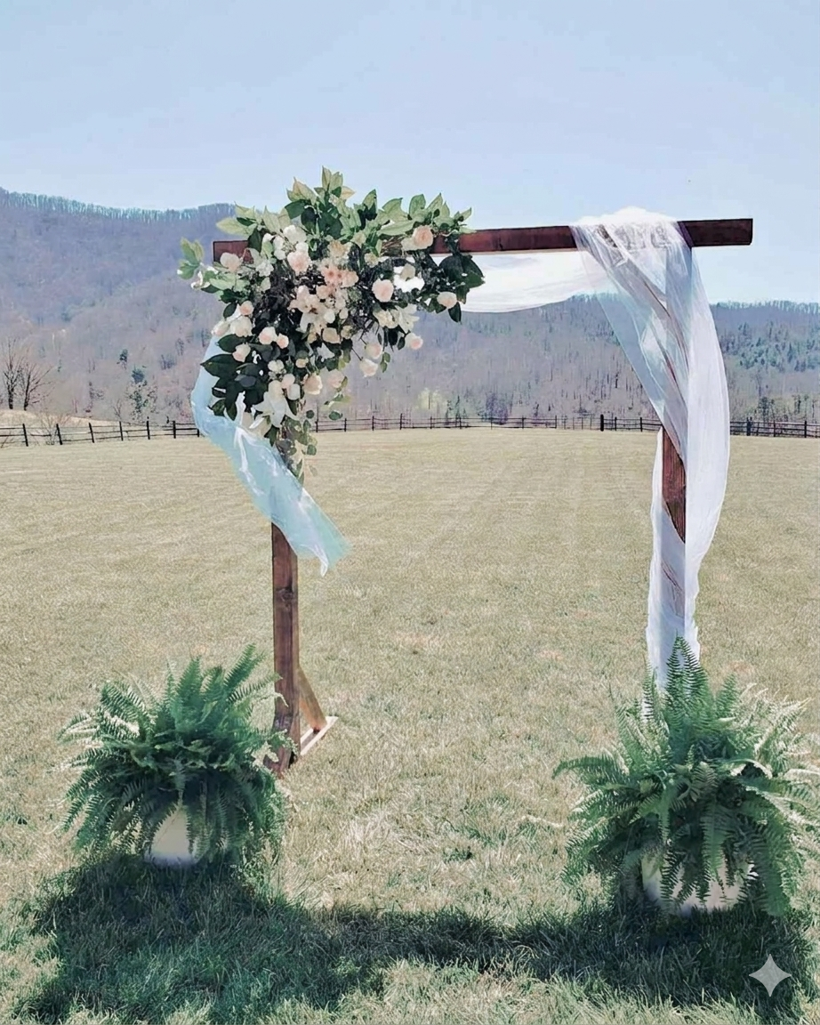 A wooden wedding arch with white and pink flowers and white fabric, standing on a grassy field with mountains in the background.