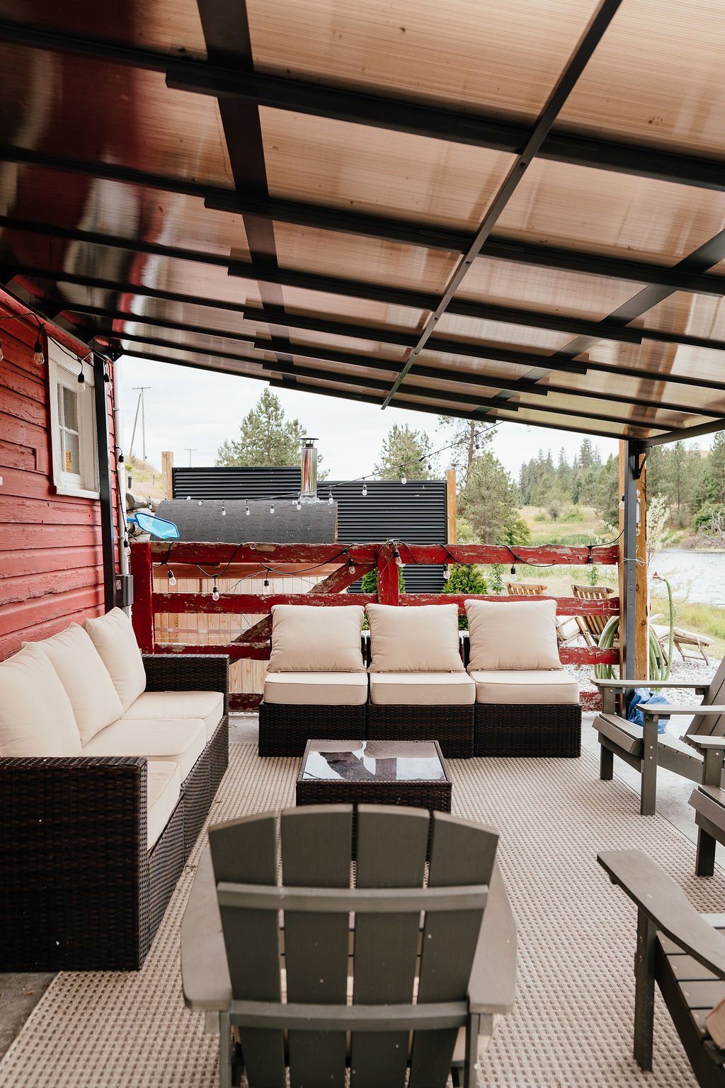 Outdoor patio with beige cushioned wicker sofas, wooden chairs, a glass coffee table, and string lights, overlooking trees and water in the background.