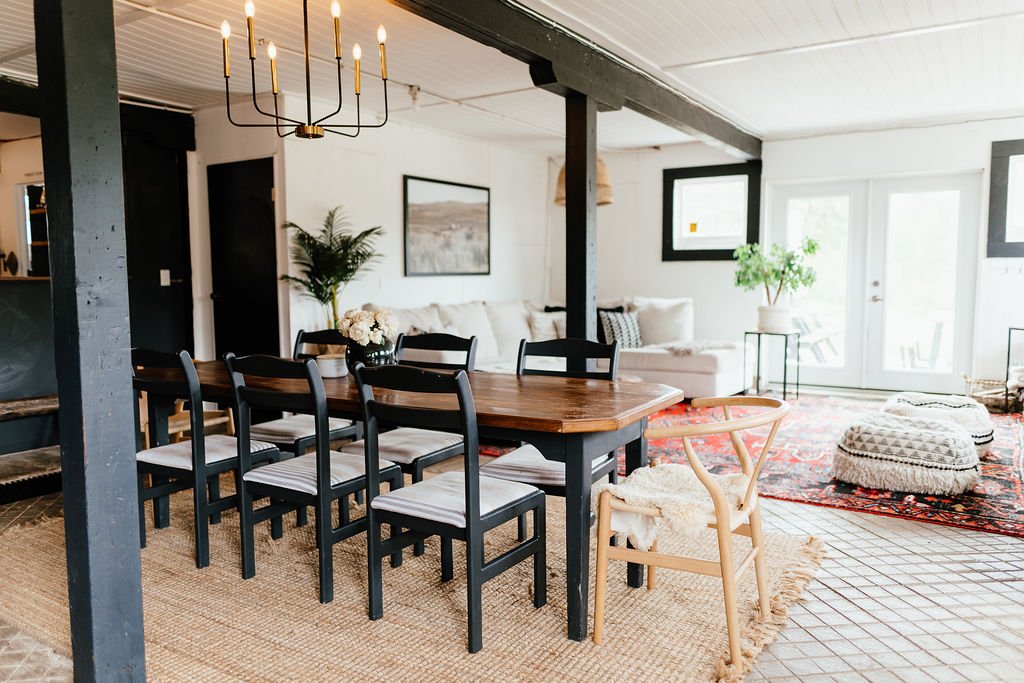 Bright living and dining room with black and wooden furniture, a chandelier, a patterned rug, and large windows with natural light.