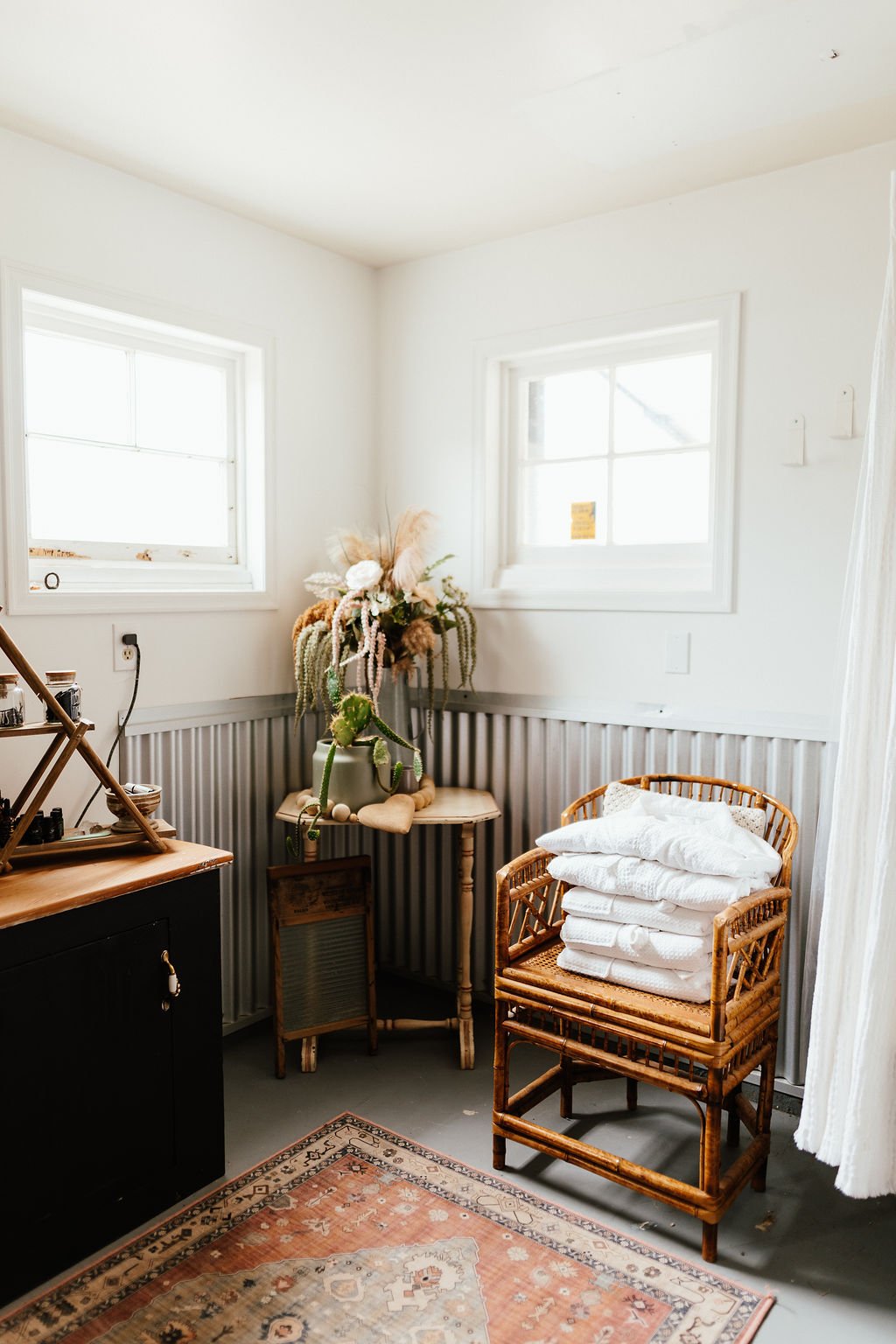 Corner of a cozy room with two small white windows, a wooden cabinet with jars on top, a black cabinet, a rug, and a wicker chair with folded white blankets. A small table with a large floral arrangement is also visible.
