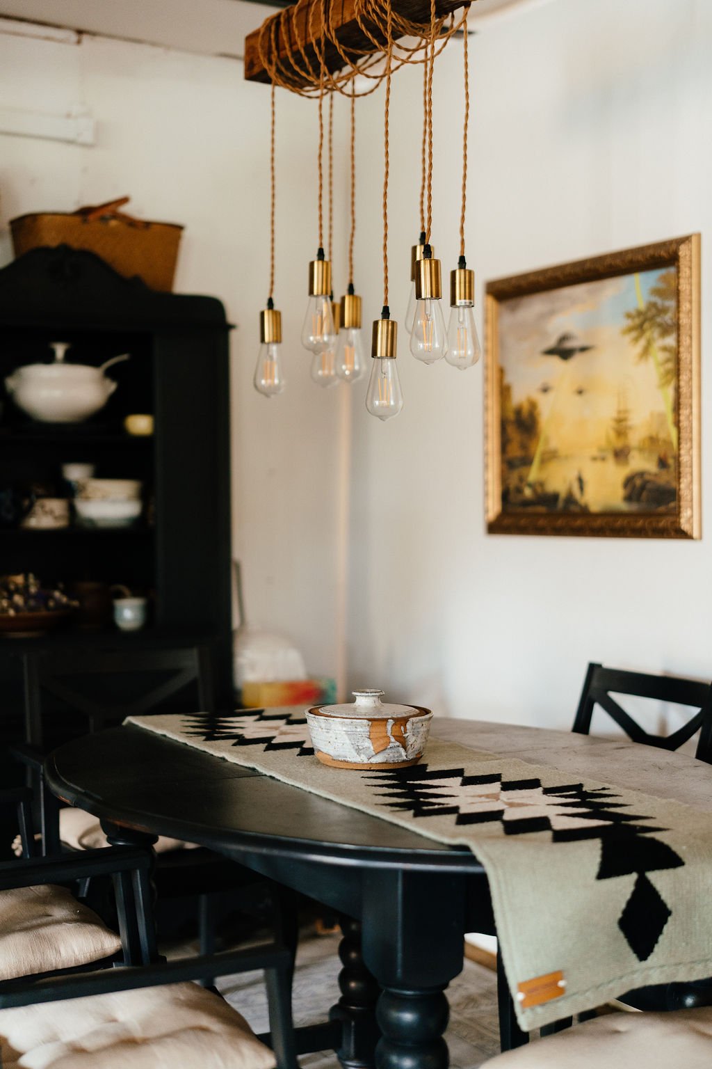 A dining room with a black table, black chairs with beige cushions, a patterned table runner, a decorative pottery piece, a black hutch with dishes, a painting on the wall, and a modern hanging light fixture with exposed bulbs and ropes.