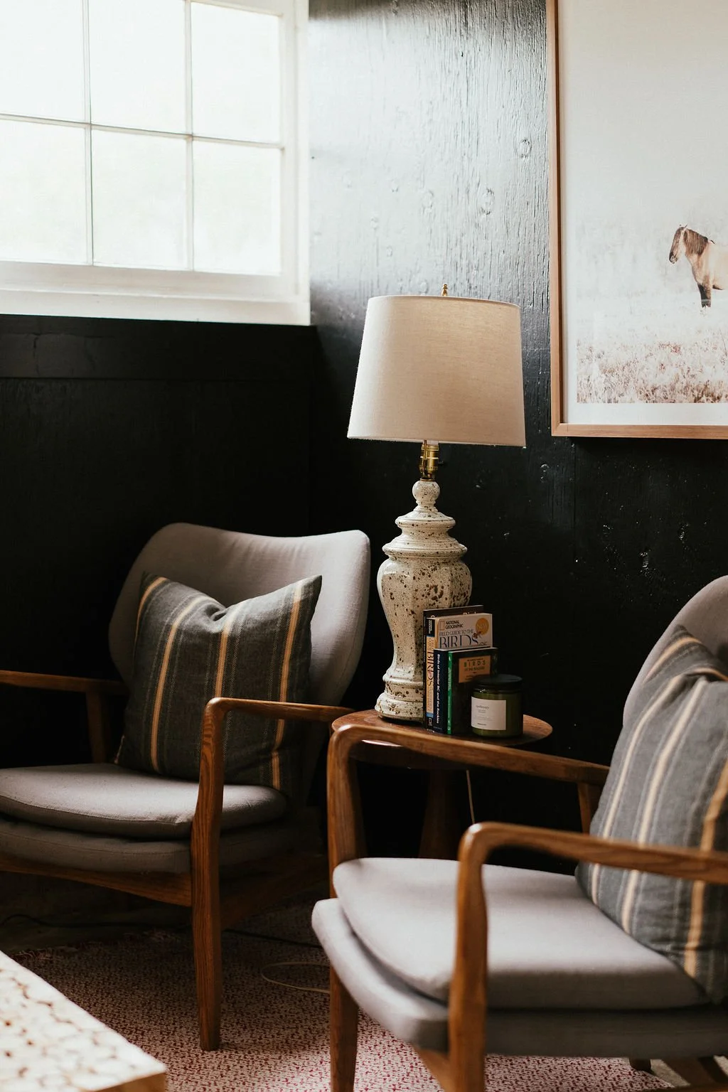 A cozy living room corner with two armchairs with plaid pillows, a wooden side table, a table lamp with a ceramic base, a few books, a small candle, and a framed picture of a horse on a textured wall.