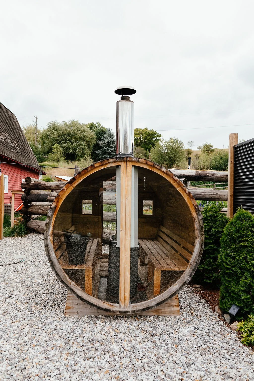 A cylindrical wooden sauna with a glass front, situated on gravel outdoors, with trees and a red house in the background.