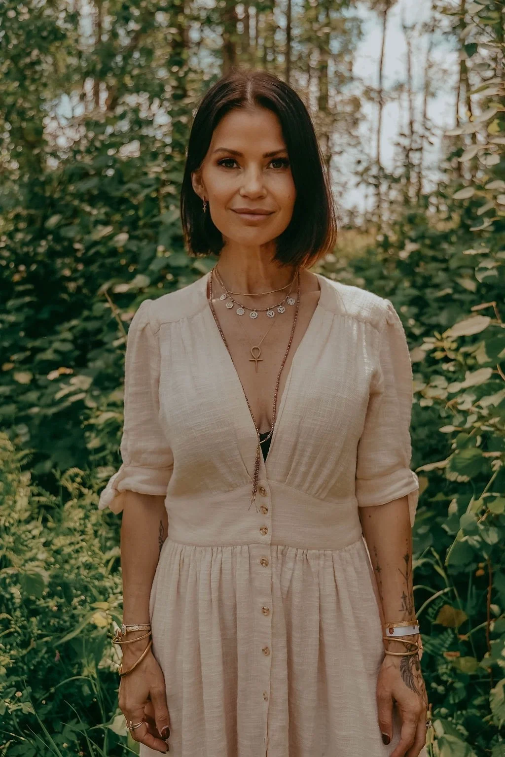 A woman with shoulder-length dark hair standing outdoors in a lush, green forest with sunlight filtering through the trees. She is wearing a light-colored, button-up dress with rolled-up sleeves, layered necklaces, and various bracelets.