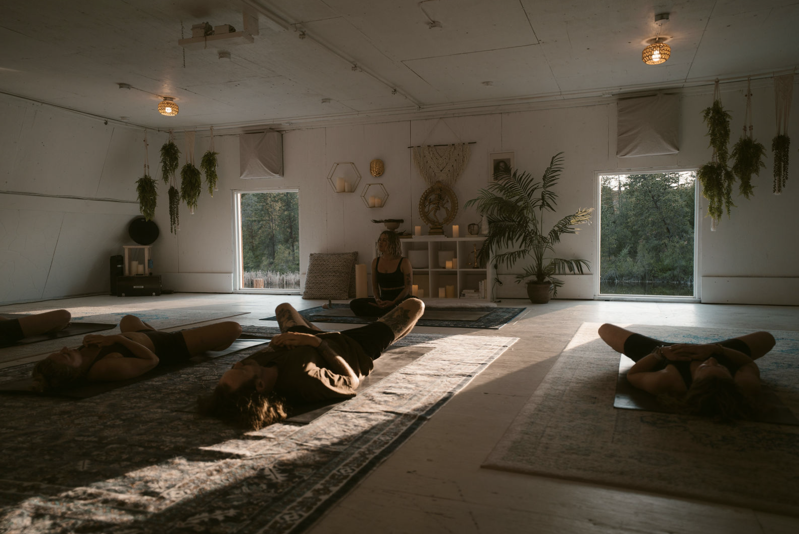 People practicing yoga in a serene, well-lit room with large windows, hanging plants, and decorative shelves with candles.