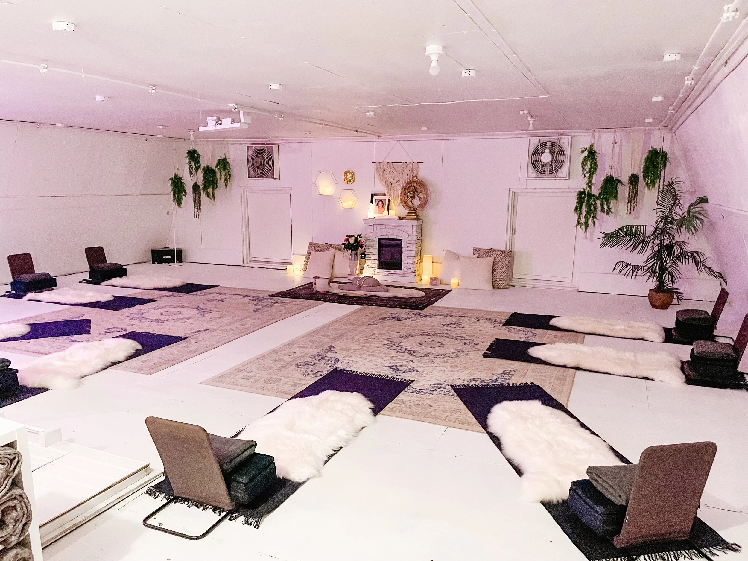 A room with a white ceiling, two square wall fans, hanging plants, a white fireplace with candles, pictures, and decorations, surrounded by cushions and small rugs on the floor, and chairs arranged around.