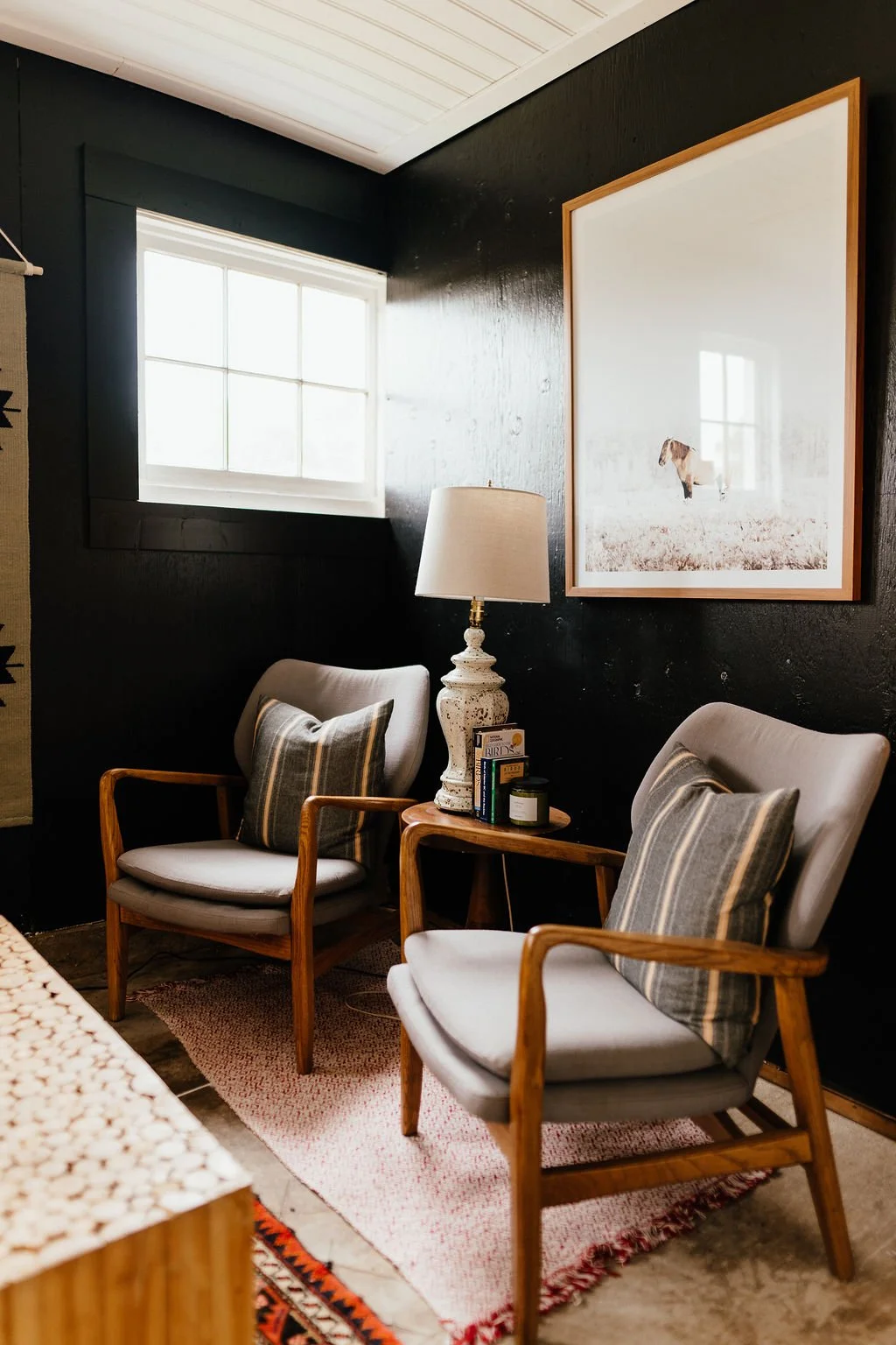 Living room with black wall, two mid-century modern armchairs with plaid pillows, a small wooden side table with a table lamp, books, and a candle, a window with white trim, and a large framed picture of a dog in a field.