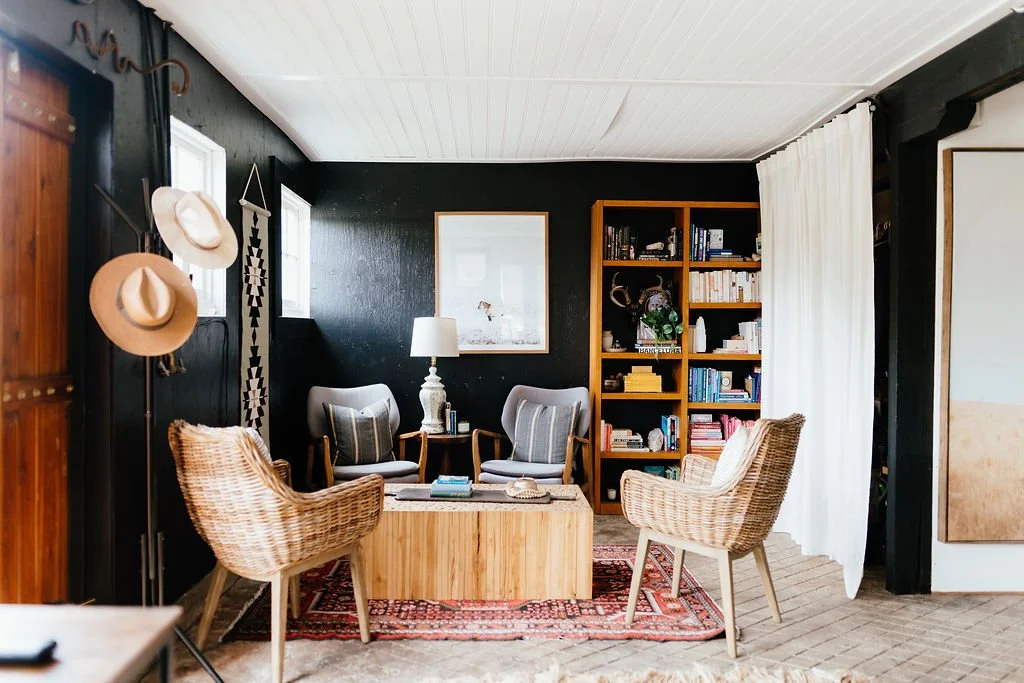 Cozy living room with black walls, wicker chairs, a wooden coffee table, a bookshelf filled with books, and a window with white curtains.