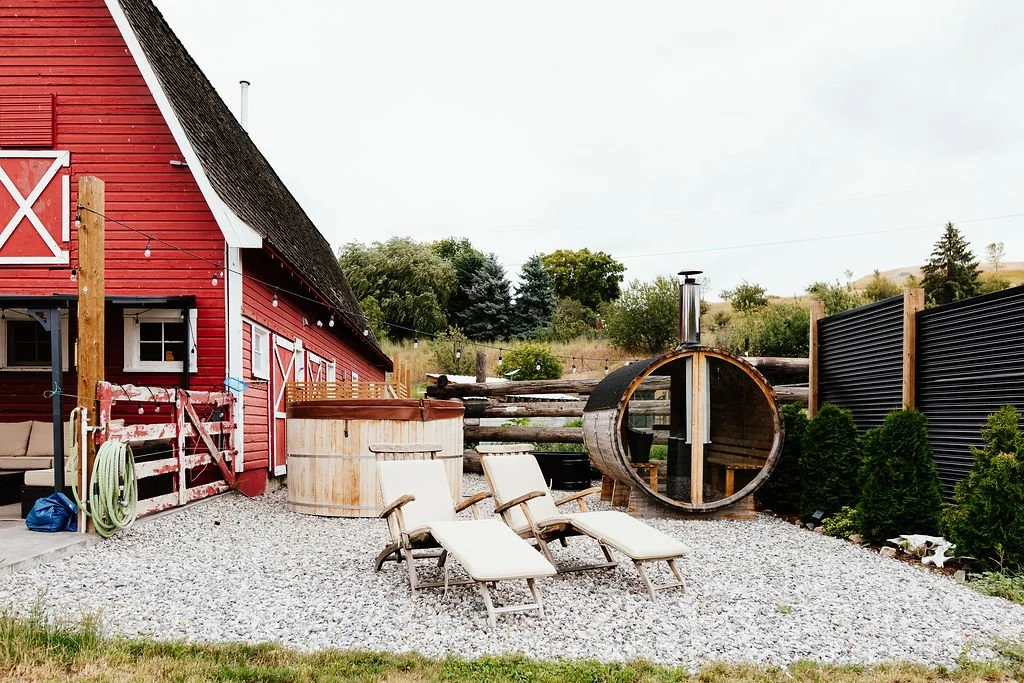 Outdoor backyard with red barn-style building, hot tub, sauna, lounge chairs, string lights, and greenery.