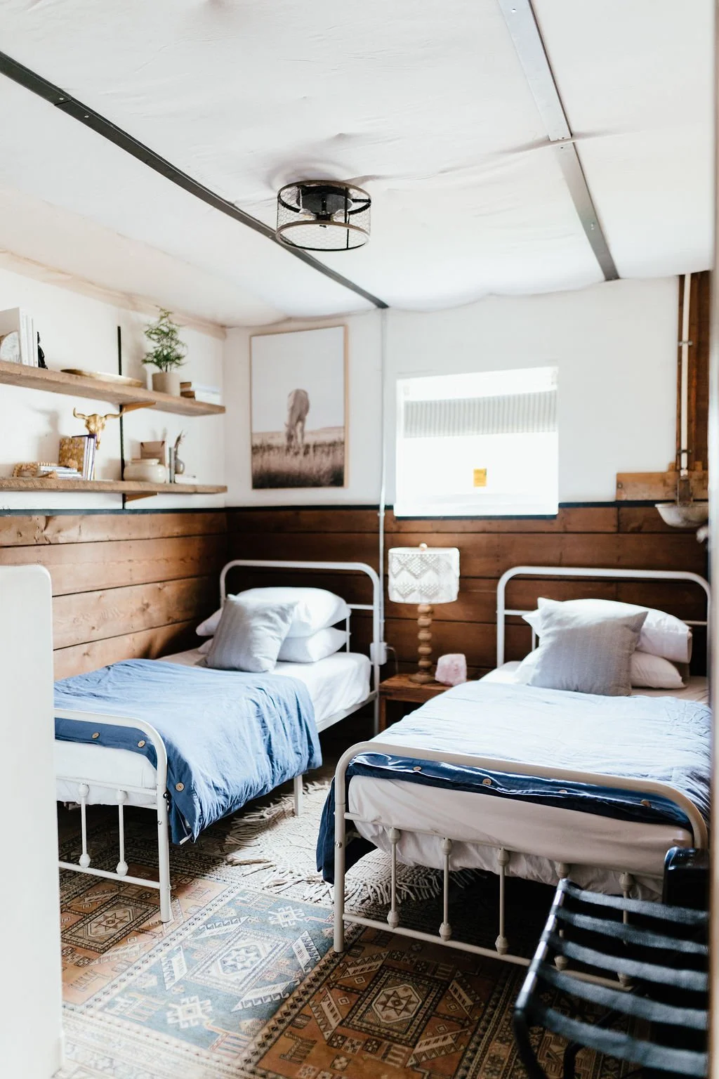 A cozy bedroom with two metal beds with white frames, pillows, and blue sheets. There's a wooden wall behind the beds, a window with blinds, a patterned rug, and a small side table with a lamp between the beds. Shelf decor and a monochrome art piece 