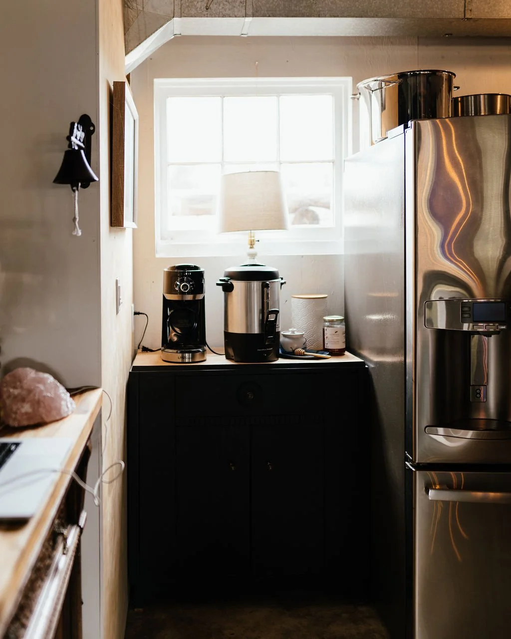 Small kitchen corner with a black cabinet, coffee maker, rice cooker, jars, and a lamp on top of a black surface near a window.