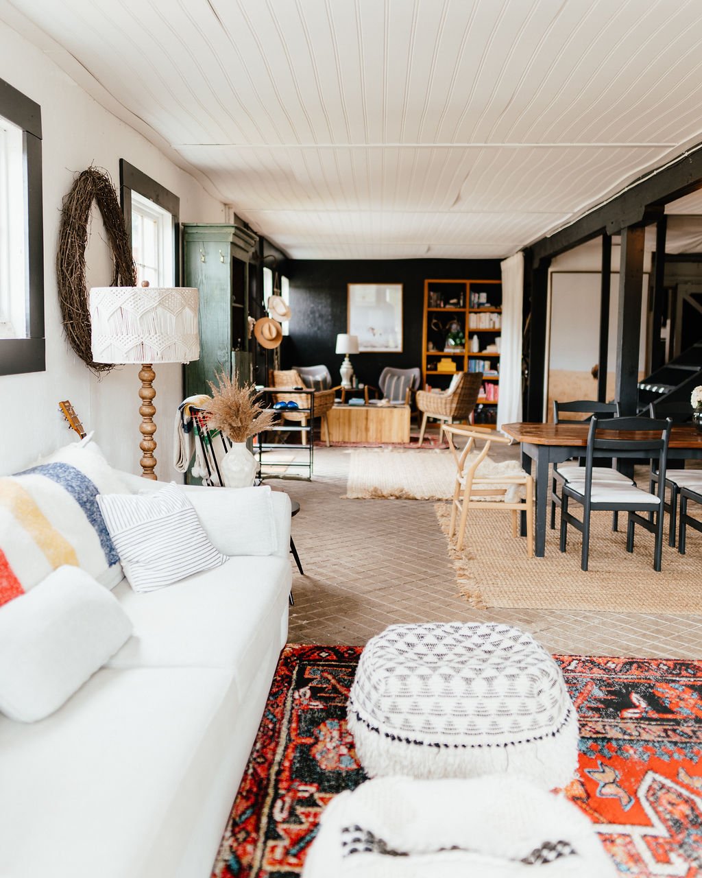 Open-concept living room and dining area with white sofa, patterned pillows, woven pouf, red and black oriental rug, wall mirror, wicker and wooden furniture, bookshelves, and decorative items.
