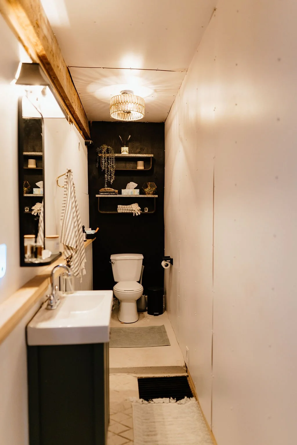 A small, modern bathroom with a white toilet, a white sink with silver fixtures, and a black wall behind the toilet. Decorative shelves with plants, candles, and ornaments are mounted on the black wall. The ceiling has a woven light fixture, and a mi