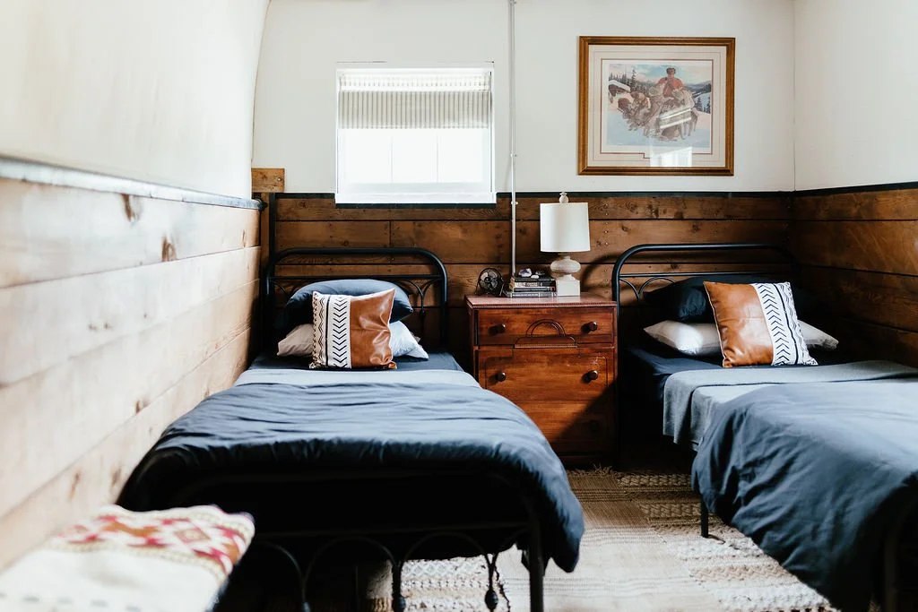 A bedroom with two single beds with black metal frames, matching pillows, and gray bedding. Between the beds is a wooden nightstand with a white lamp, books, and a vintage phone. A framed picture hangs above. A small window with blinds is on the wall.