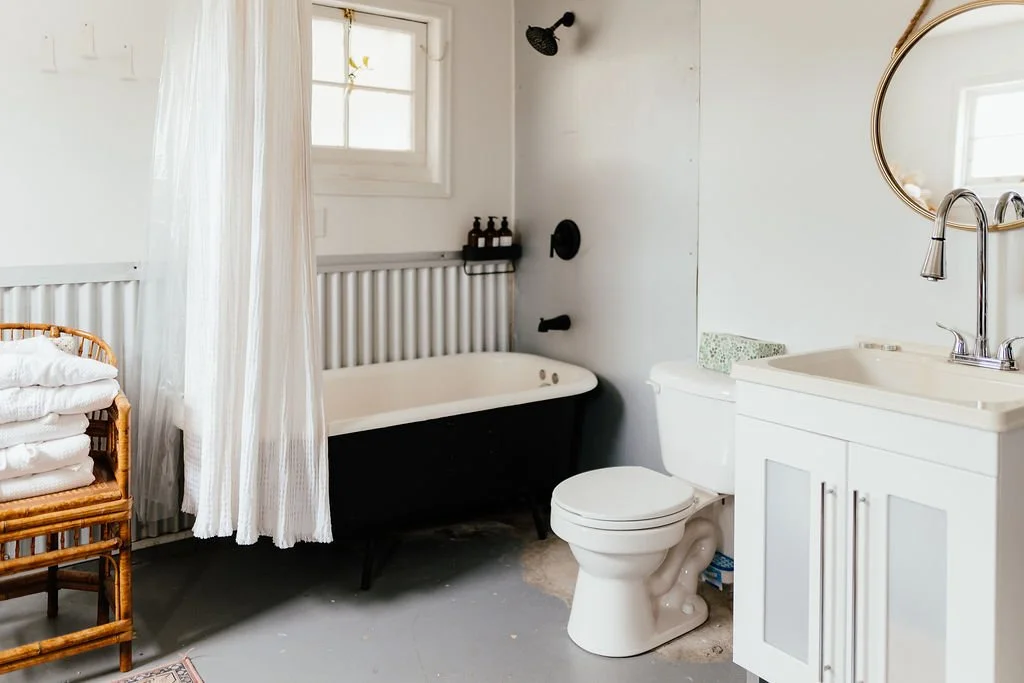A bathroom with a bathtub next to a window, a toilet, a sink with a mirror, a wicker basket with towels, and some bottles of soap or shampoo.