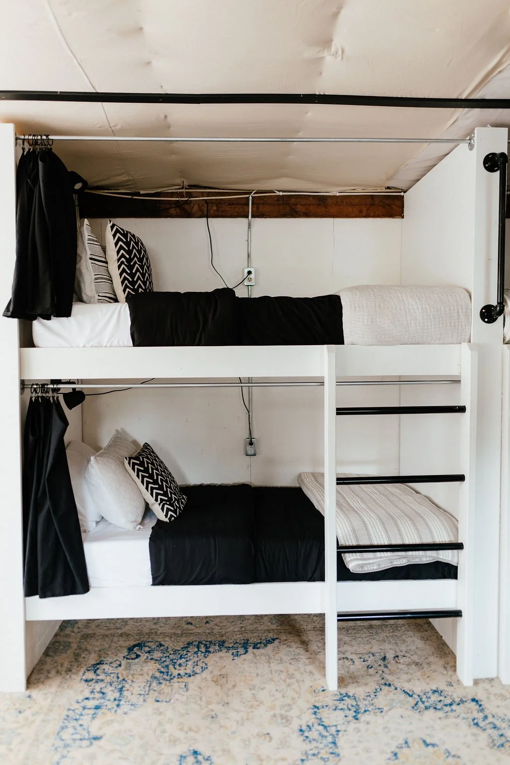 Bunk beds with black and white bedding and patterned pillows, black curtains on the sides, and wooden beams on the ceiling, in a room with a beige rug and white walls.