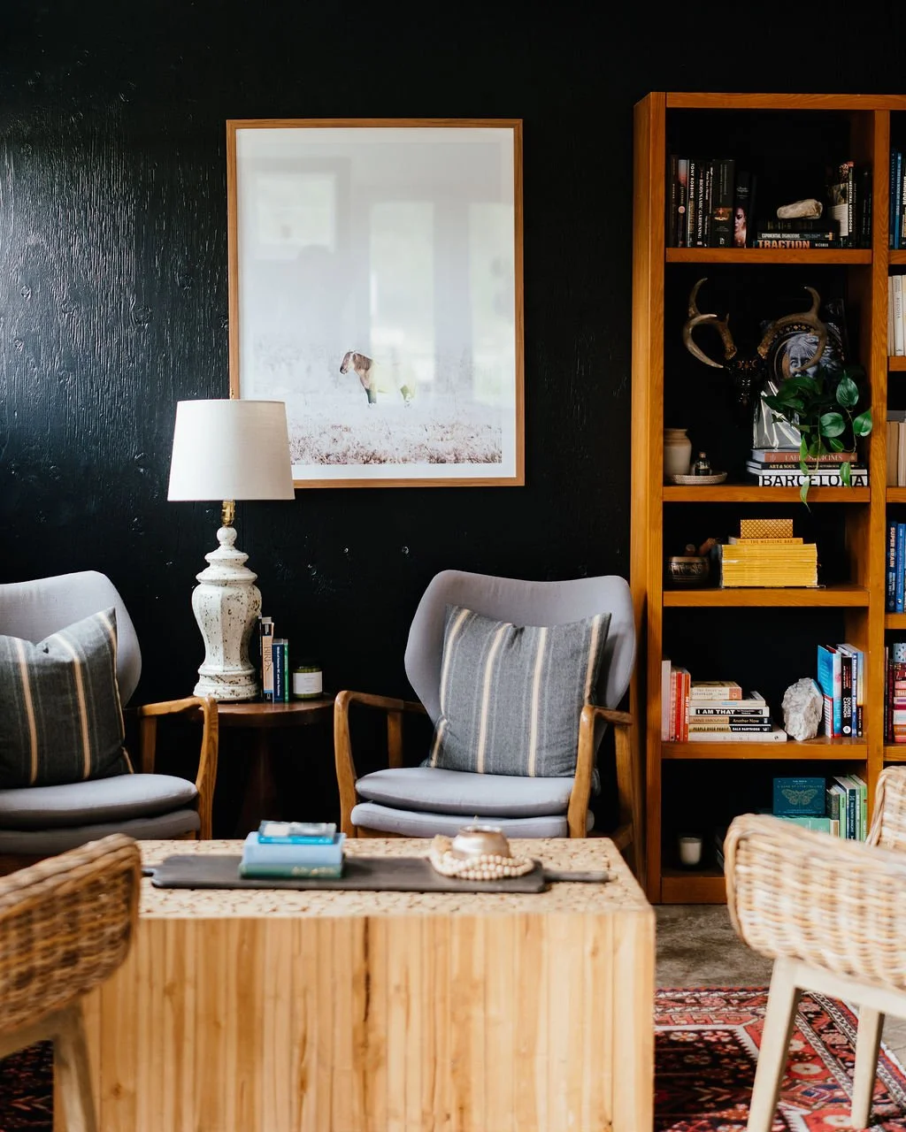 Living room with two grey armchairs, a white table lamp on a side table, a wooden bookshelf filled with books and decorative items, a framed photograph of a dog on the dark wall, and a wooden coffee table with books and decorative objects.