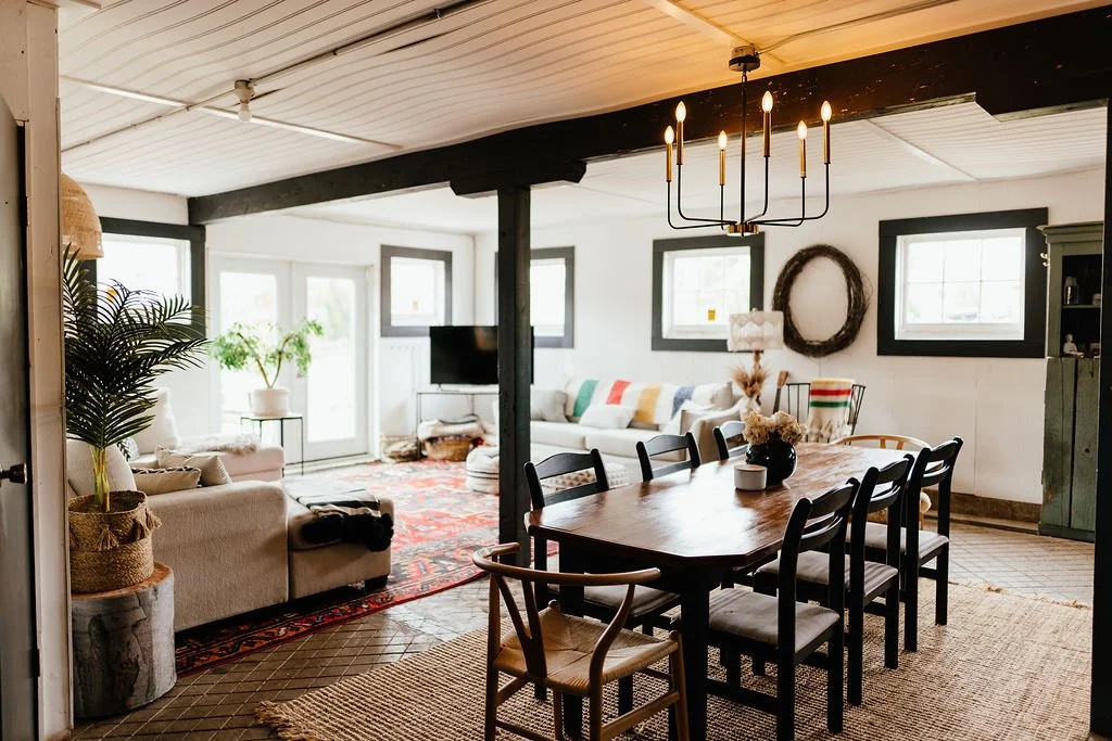Living and dining room with white walls, black window frames, a black chandelier, and various furniture including sofas, a dining table with chairs, and a TV.