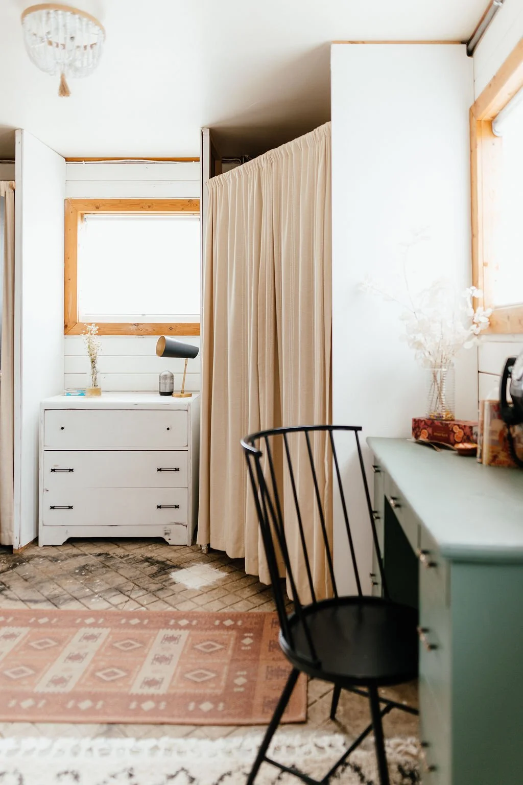 Interior of a cozy, minimalist room with white walls, wooden trim, a white dresser, black chair, a small desk, beige curtains, and decorative vases with dried flowers.
