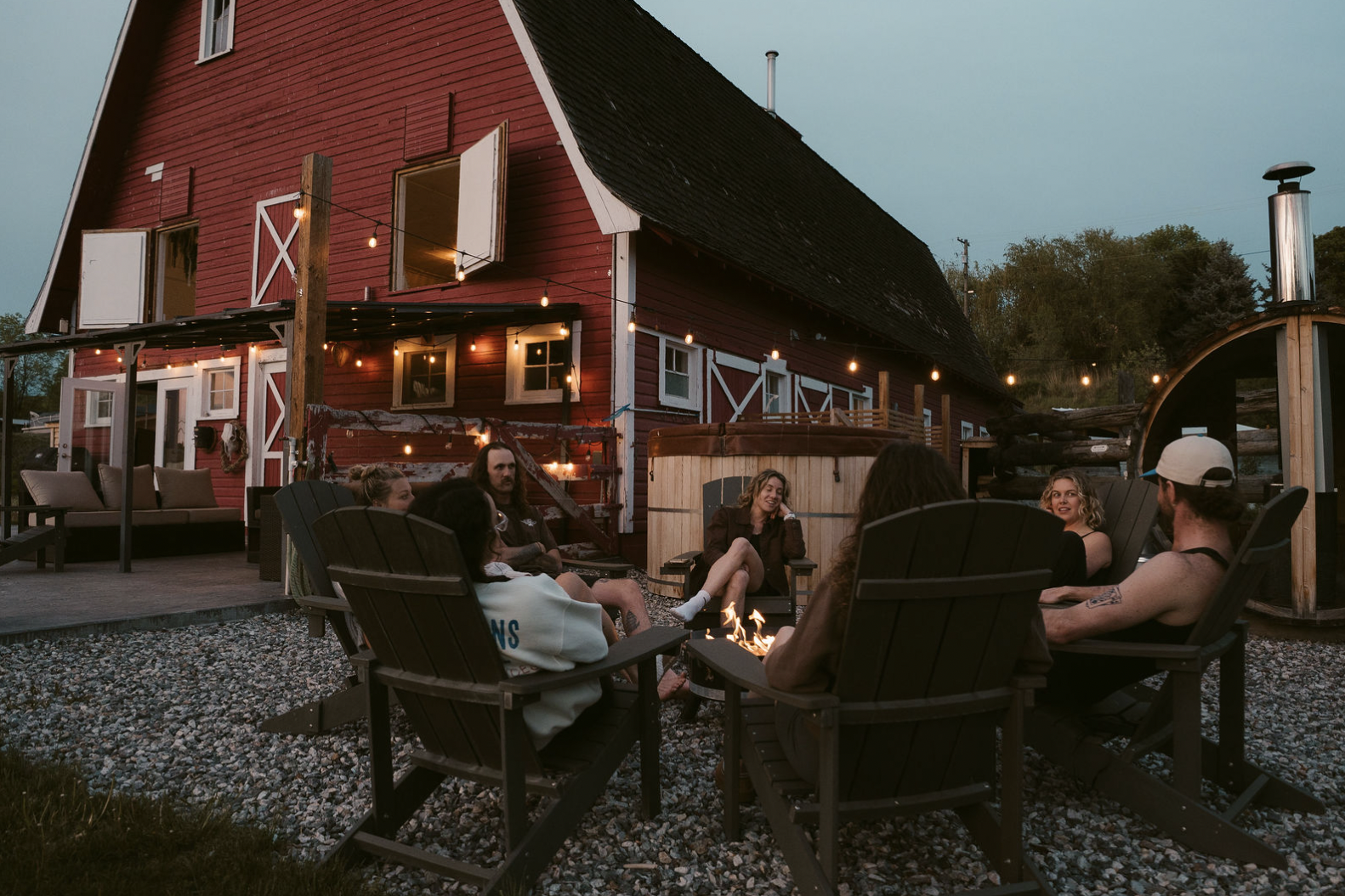 Group of people sitting around a firepit outdoors at dusk in front of a red barn with string lights, log pile, hot tub, and outdoor furniture.