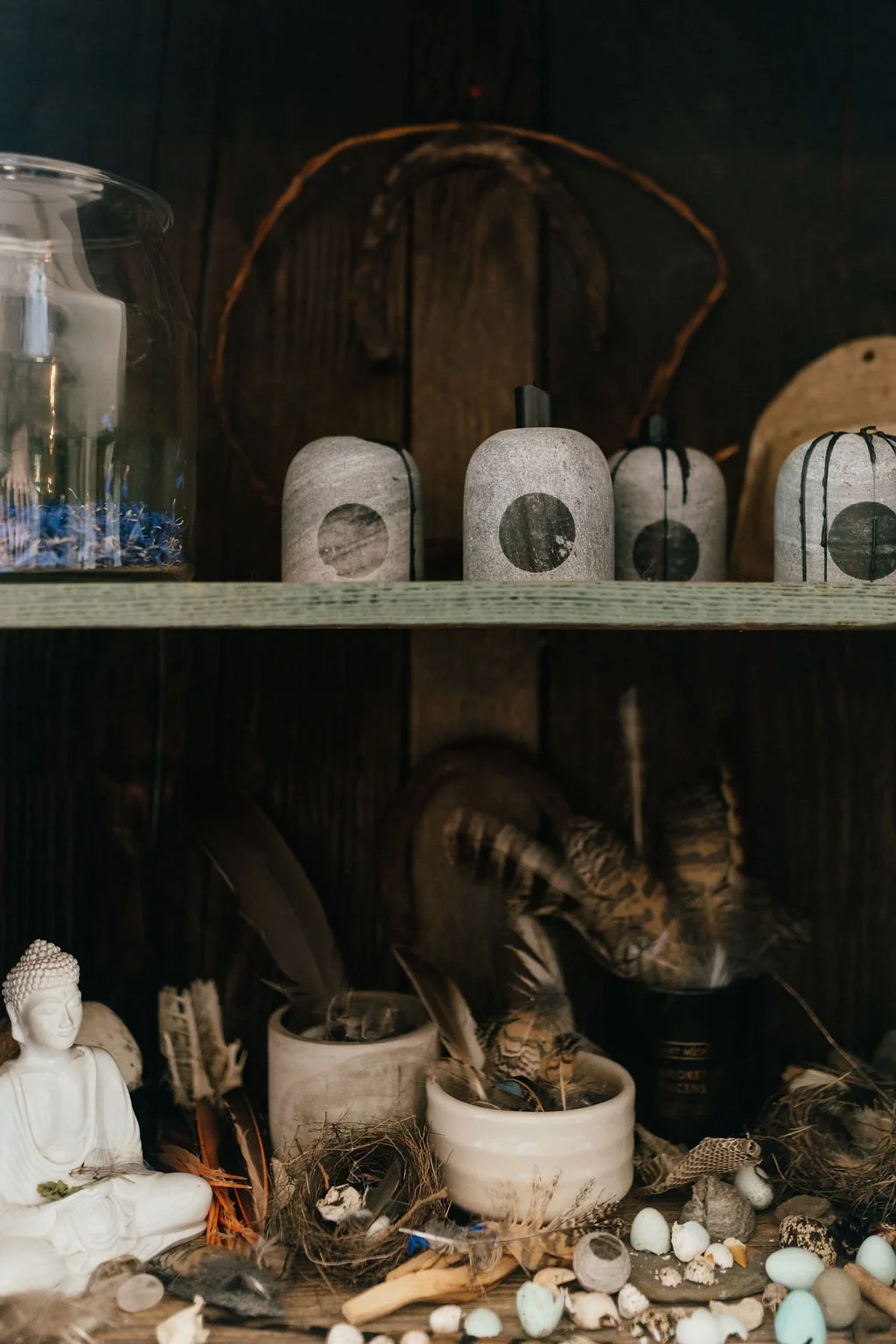 Shelf displays decorative items including a small white Buddha statue, peacock feathers in containers, and a jar of blue feathers, with scattered eggs and natural materials around.