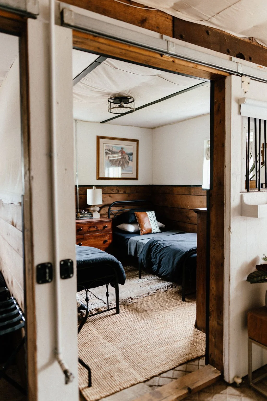 A rustic bedroom with two beds, dark bedding, a wooden nightstand with a lamp, a framed picture on the wall, and a woven rug on the floor.