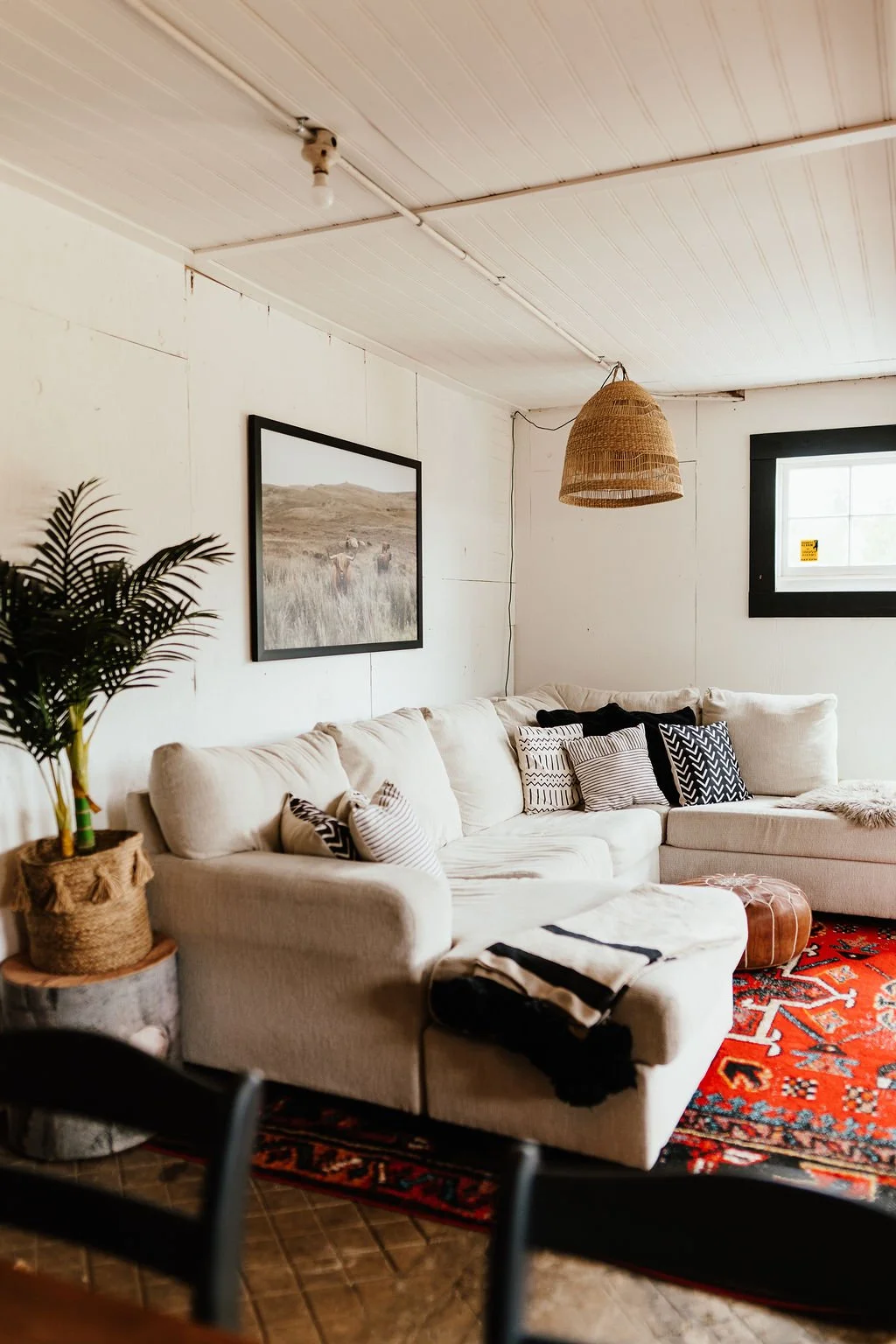 Living room with white sectional sofa, black and white patterned pillows, a red patterned area rug, a wicker pendant light, a potted plant, wall art, and a small window with black trim.