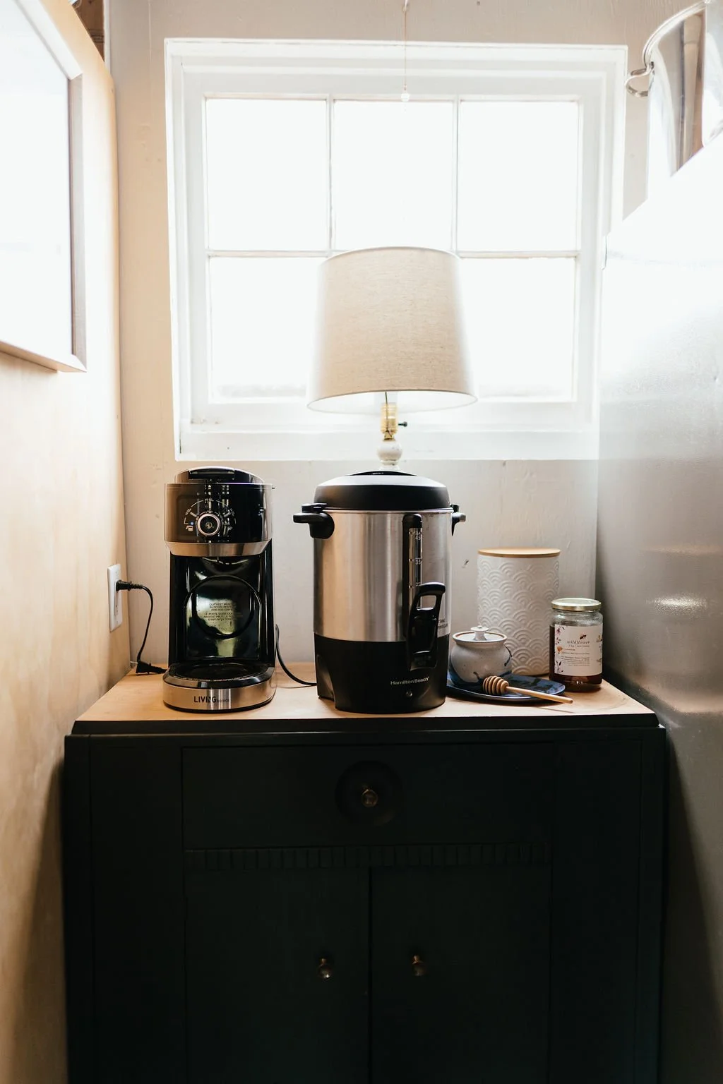 Coffee maker, rice cooker, and other kitchen items on a wooden table against a window with a lamp on it.