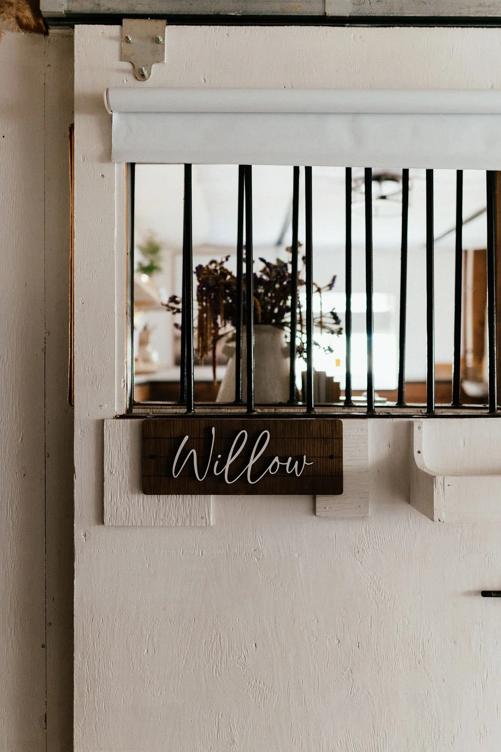 A wooden sign with the name 'Willow' hangs below a window with black bars. Behind the window, there is a vase with dried flowers on a table in a cozy, decorated interior.