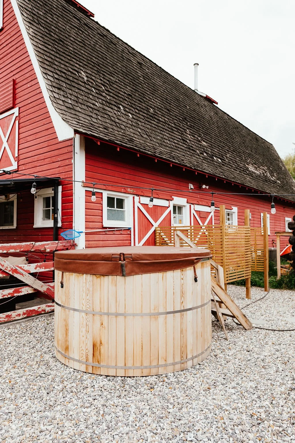 A red barn with white trim and small windows, outdoor string lights, a wooden hot tub with a brown cover, and gravel ground.