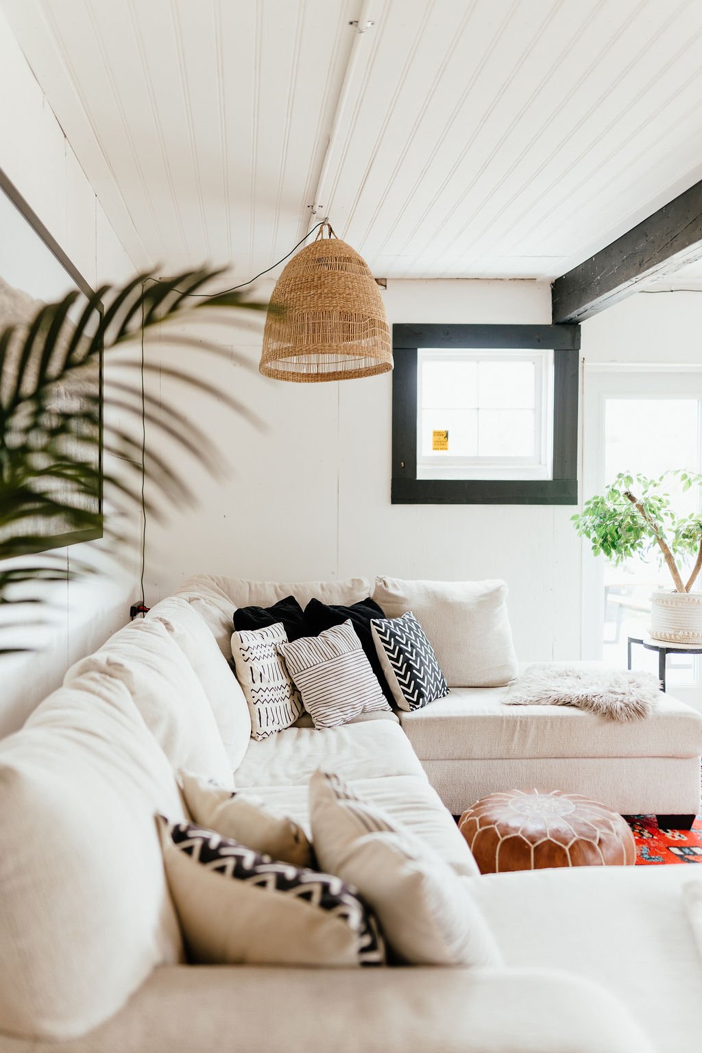 A living room with a cream-colored sectional sofa decorated with black and white patterned pillows, a woven hanging light fixture, a potted plant on a side table, and natural light coming through a window.