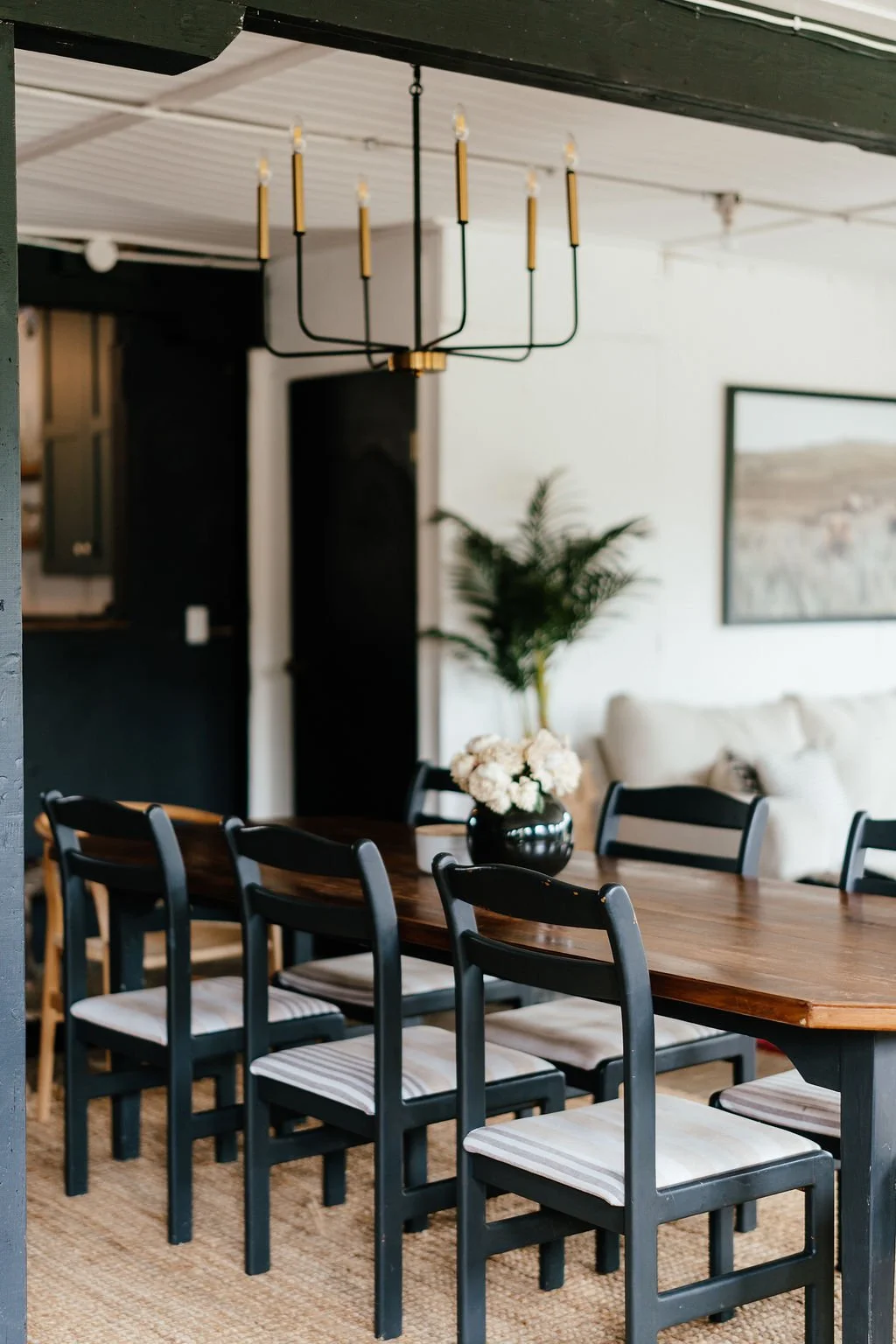 A dining room with a dark wooden table, black chairs with striped cushions, a black vase with white flowers, and a large plant in the background, with a framed landscape painting on the white wall and a modern chandelier hanging from the ceiling.